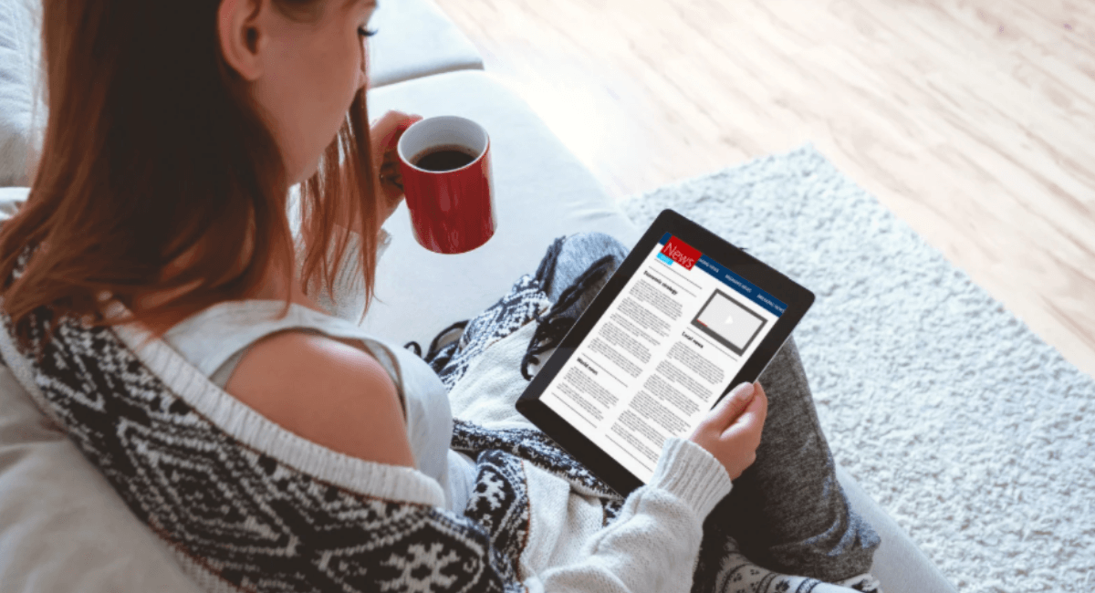 A woman relaxing on a couch, holding a red coffee mug in one hand while reading a news website on a tablet in the other.