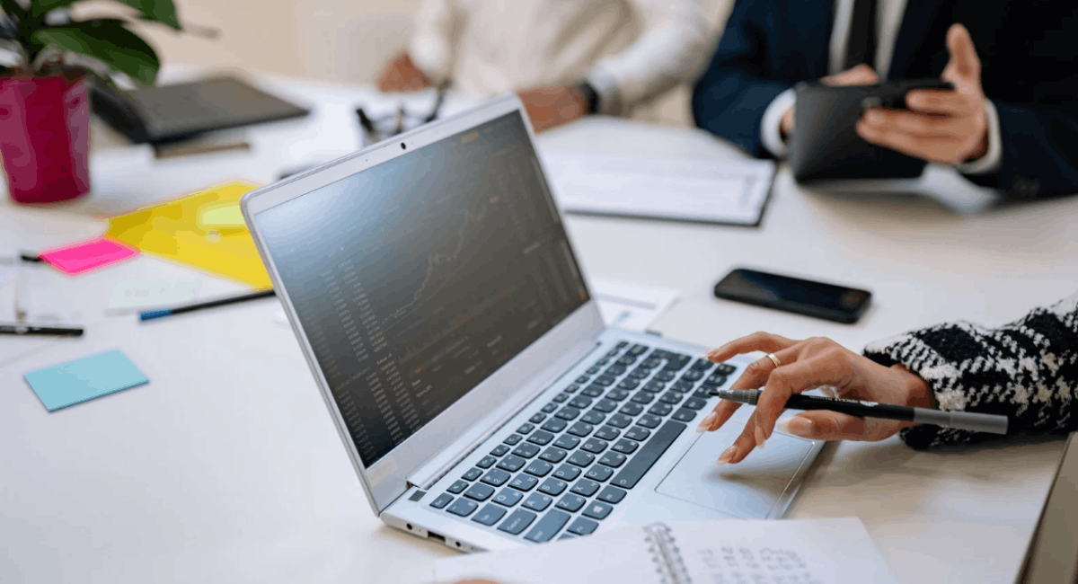 Close-up of a person’s hand using a laptop trackpad at a meeting table, with data charts visible on the screen and colleagues in the background.