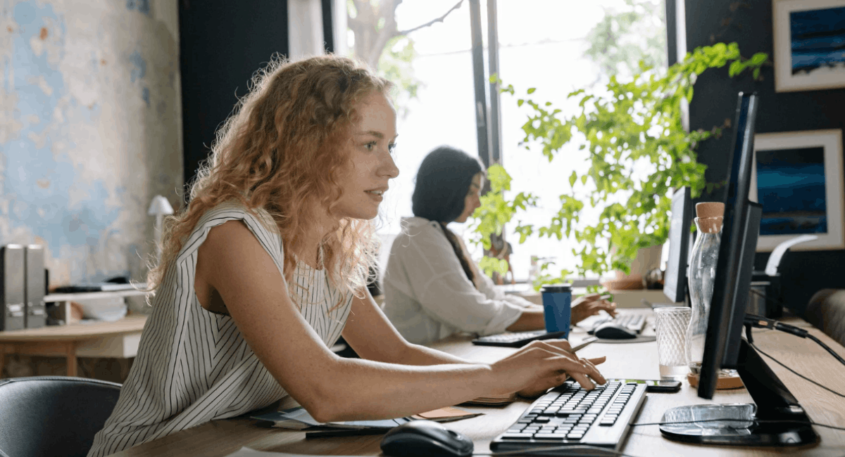 A woman with curly hair sitting at a desk in a bright office, typing on a keyboard while focused on her computer monitor.