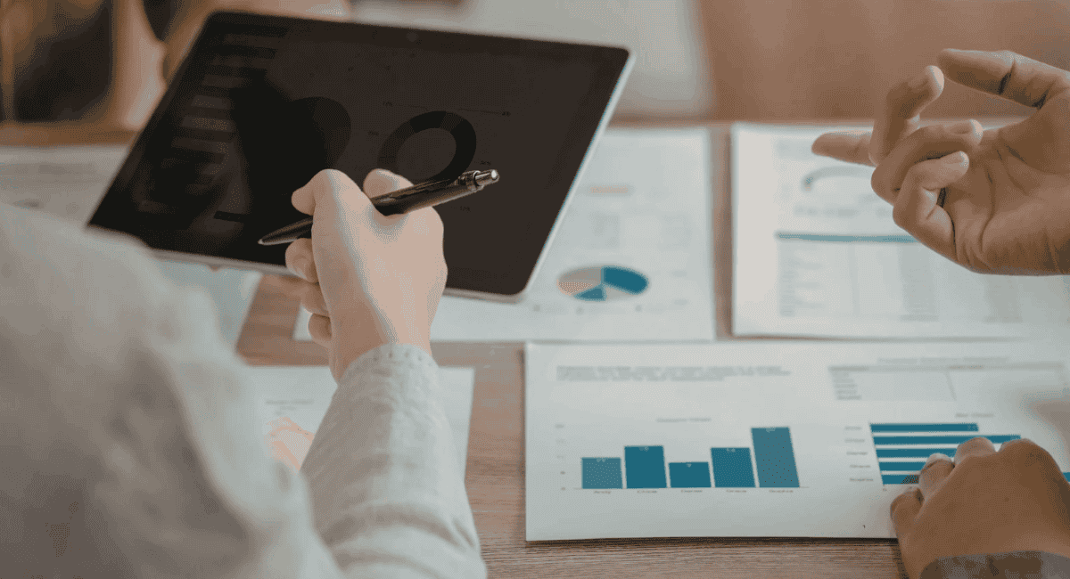 Two people in a meeting reviewing data, with one person holding a tablet displaying a pie chart and another pointing at printed bar graphs on the desk.