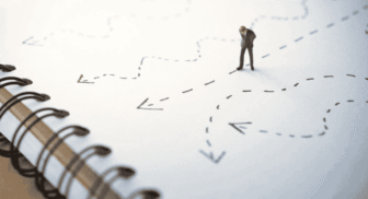 A miniature figurine of a businessman standing on a notebook surrounded by hand-drawn dashed lines and arrows pointing in various directions.