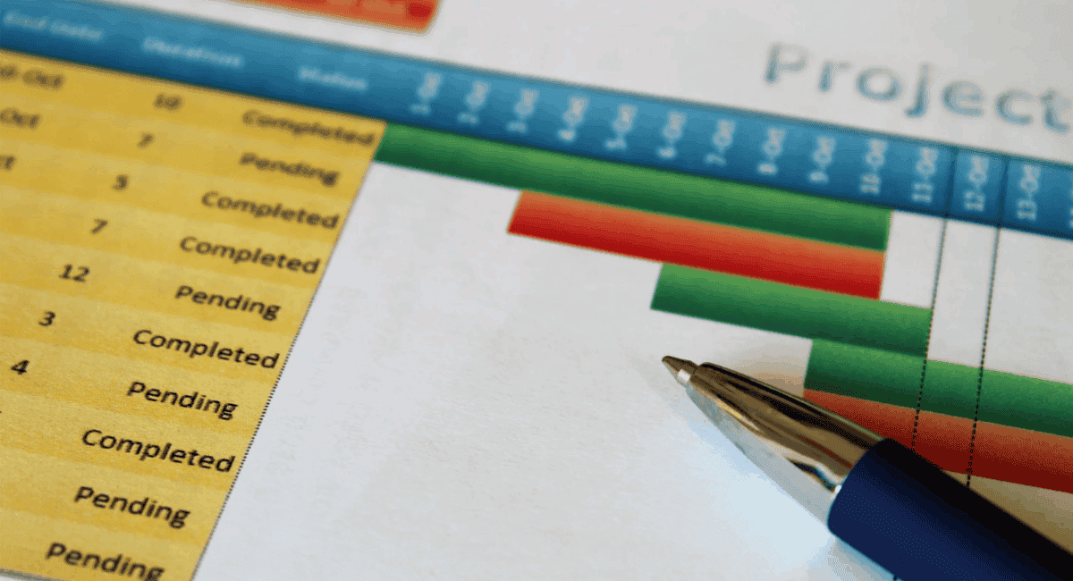 A close-up of a printed project management Gantt chart with status columns and color-coded timeline bars, with a blue pen resting on the paper.