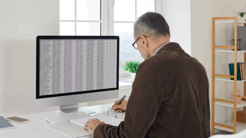 A man from a back-view perspective sitting at a white desk, analyzing a massive data spreadsheet on a large monitor and taking notes.