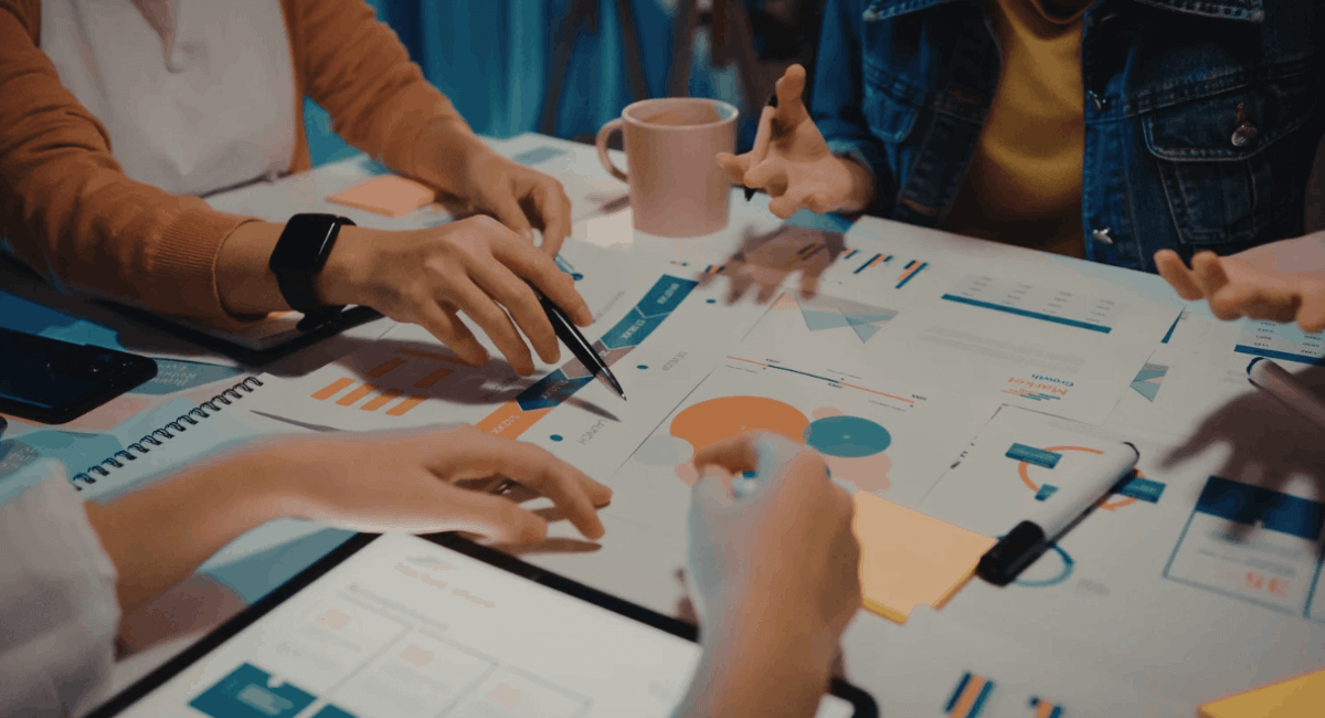 A close-up view of a team's hands analyzing printed marketing graphs, charts, and sticky notes spread across a conference table.
