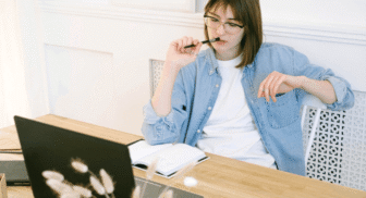 A young woman with glasses and a denim shirt sitting at a wooden desk. She is holding a pen to her lips in a thoughtful pose, with an open notebook and a laptop in front of her.