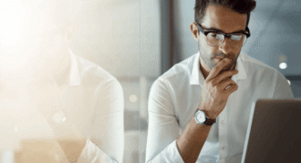 A focused professional wearing glasses looking down intently at his laptop screen, with his reflection visible in a glass partition next to him.