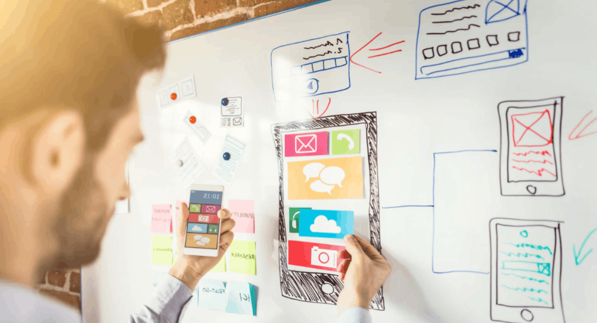 A web developer planning a mobile user interface by sticking colored UI element cutouts onto a smartphone wireframe drawn on a whiteboard.