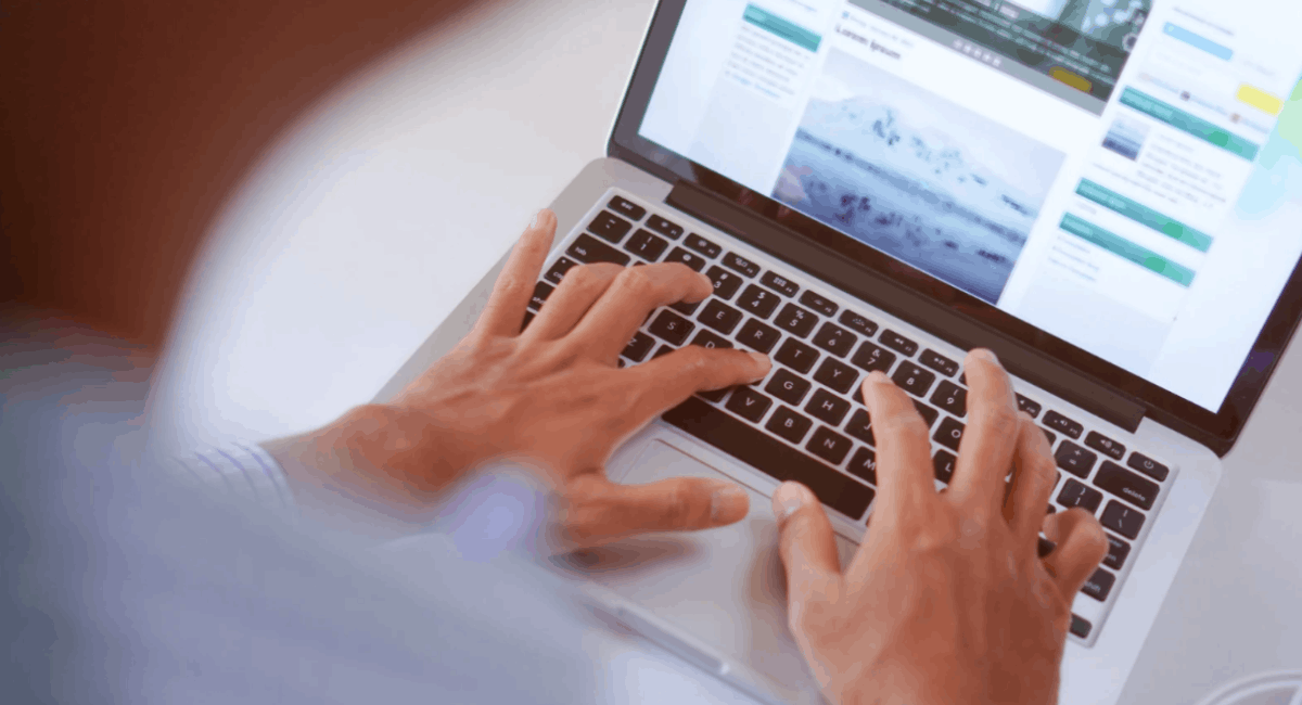 A high-angle, close-up shot of a person’s hands typing on a silver laptop keyboard. The screen shows a webpage with a light-colored layout and images.