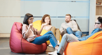 A group of four colleagues wearing casual clothing sitting in colorful beanbag chairs and engaging in a collaborative team discussion.