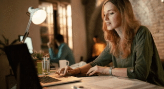 A woman sitting at a desk under a desk lamp at night, focused on typing on her laptop while holding her smartphone.