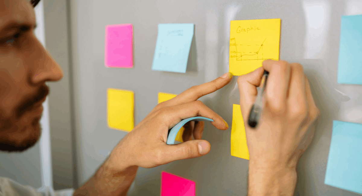 Side profile of a man with a beard pointing at a yellow sticky note on a glass wall while organizing a colorful grid of post-it notes during a planning session.