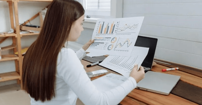 The Always Up-to-Date SEO Checklist插图13 Over-the-shoulder view of a woman with long brown hair sitting at a wooden desk, comparing printed business reports featuring colorful bar charts and line graphs next to an open laptop.