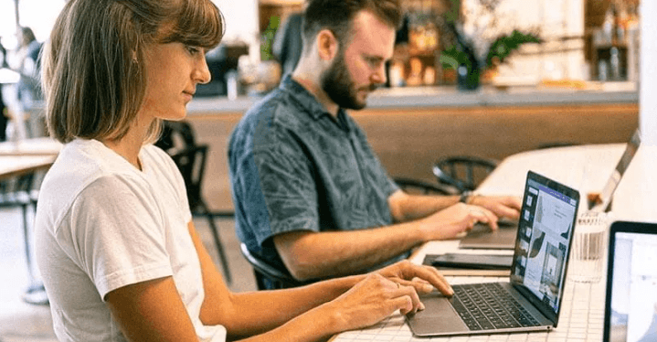 The Always Up-to-Date SEO Checklist插图8 A woman and a man sitting side-by-side at a long white counter, working independently on their laptops in a modern office or coffee shop environment.
