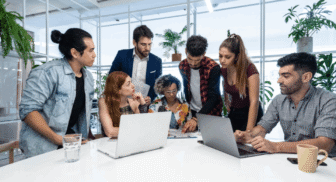 A diverse group of seven colleagues gathered around a white office table, collaborating closely while looking at laptops, documents, and discussing a project.
