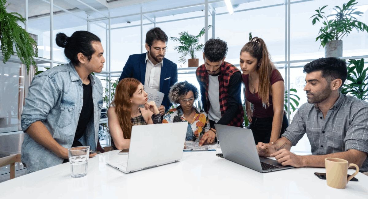 A diverse group of seven colleagues gathered around a white office table, collaborating closely while looking at laptops, documents, and discussing a project.