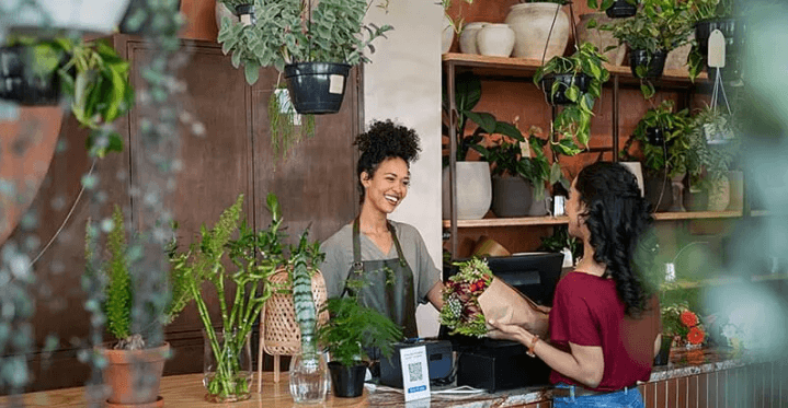 The Always Up-to-Date SEO Checklist插图5 A smiling florist behind a wooden shop counter handing a wrapped bouquet of flowers to a customer at checkout in a plant-filled boutique.