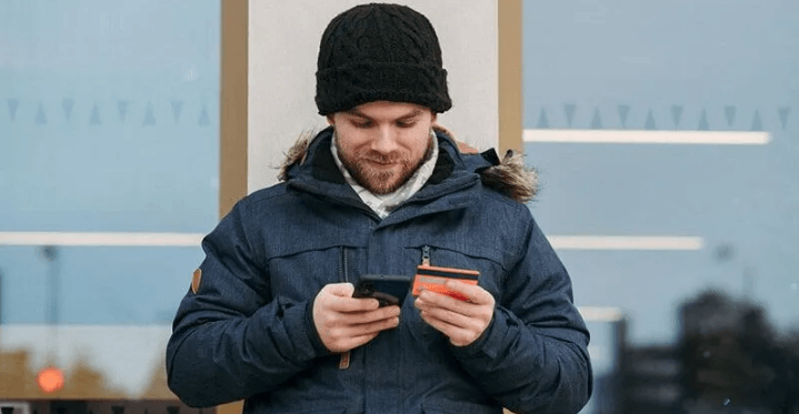 The Always Up-to-Date SEO Checklist插图6 A man wearing a winter beanie and jacket standing outdoors while holding a smartphone in one hand and a credit card in the other to complete a purchase.