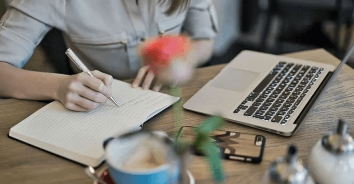 The Always Up-to-Date SEO Checklist插图1 Close-up of a person writing in a notebook with a pen at a wooden table, with an open laptop, smartphone, and a blurry coffee cup nearby.
