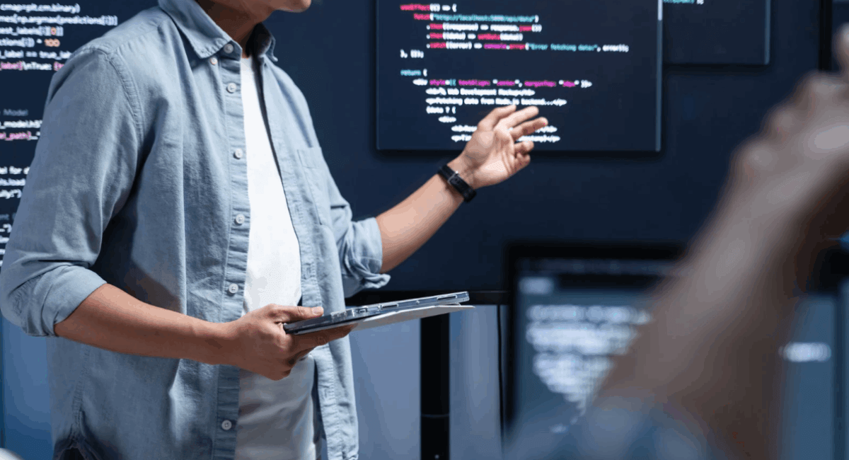 Midsection of a software developer in a blue button-down shirt holding a tablet and gesturing towards a large monitor displaying blocks of code during a technical presentation.