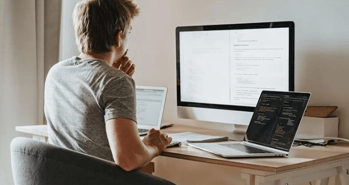 The Always Up-to-Date SEO Checklist插图4 A man sitting in front of a large desktop monitor and two open laptops, focused on writing and analyzing code at his home office desk.