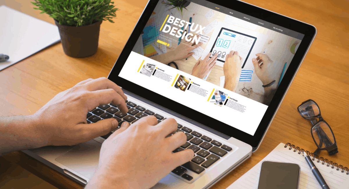Top-down view of a person working on a laptop displaying a webpage layout titled 'BEST UX DESIGN' on a clean wooden desk, next to a notebook, smartphone, glasses, and a small potted plant.