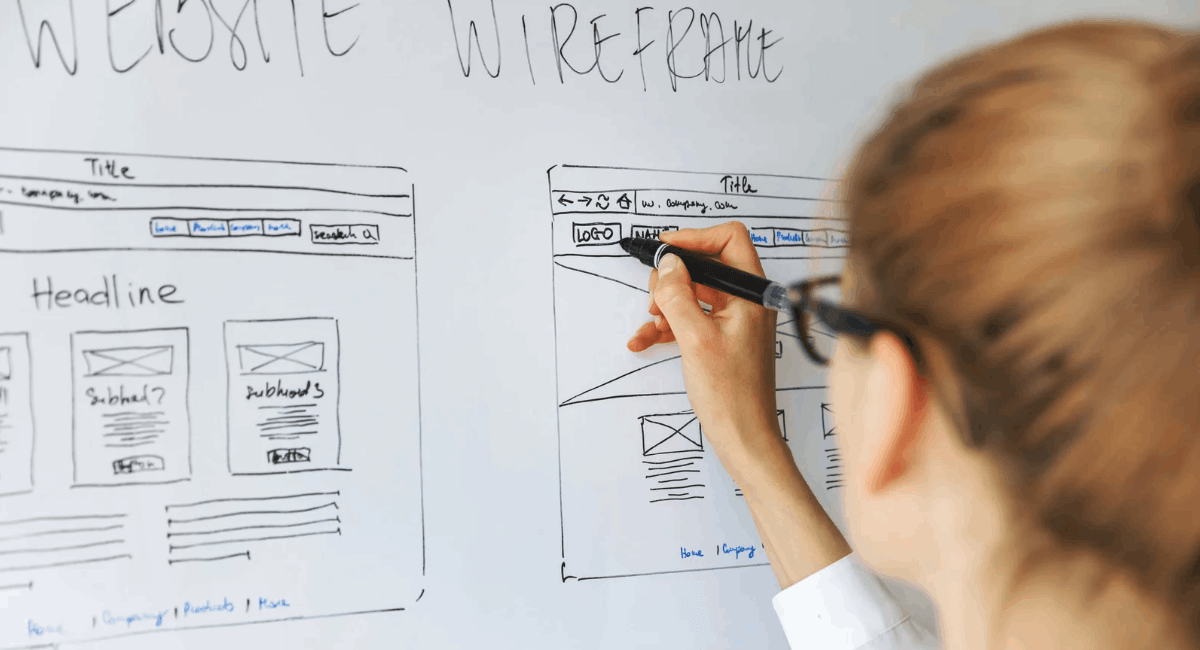 A close-up over-the-shoulder view of a woman with glasses drawing a website wireframe layout on a whiteboard with a black marker.