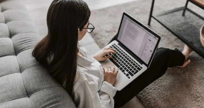 The Always Up-to-Date SEO Checklist插图2 Overhead view of a woman with glasses sitting on a rug and leaning against a grey couch while typing a document on her laptop.